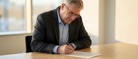 Mature male professional with gray hair and glasses wearing dark blazer and light blue shirt, writing on document at wooden desk in modern office with window background.