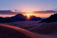 Dramatic sunset over sand dunes creates golden light rays piercing through mountain silhouette, casting long shadows across desert landscape in purple and orange hues.
