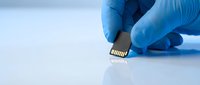 Hand in blue latex glove holding a micro SD memory card over reflective white surface, symbolizing data storage and technology.