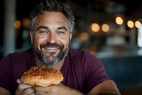 Mature man with gray beard wearing purple t-shirt smiling while holding fresh burger in casual restaurant setting with warm bokeh lights in background.