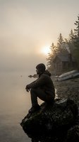 Solitary man in winter clothing sits on rocky shore contemplating misty lake at sunrise with rustic cabin and evergreen trees in atmospheric morning light.