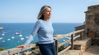 Mature woman enjoying coastal vacation standing on stone terrace overlooking Mediterranean sea with boats. Relaxed retirement lifestyle concept.