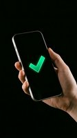 Hand holding smartphone displaying glowing green checkmark on black screen, symbolizing approval, success, and confirmation for app development, user interface, and digital validation concepts.