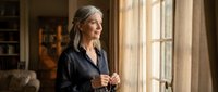 Mature businesswoman with gray hair standing by window in modern office, contemplating future plans and career decisions in warm natural light.