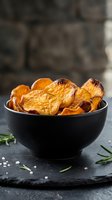 Crispy baked sweet potato chips in black bowl with rosemary herbs and sea salt on dark slate surface for healthy snacking and cooking concepts.