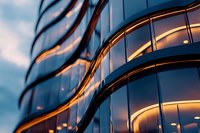 Modern curved glass building facade with warm evening light reflecting on windows creating abstract geometric pattern against blue twilight sky.