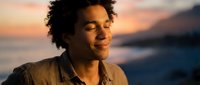 Young mixed-race man with curly hair smiling peacefully with eyes closed during golden hour sunset by ocean. Serene moment of happiness and relaxation.