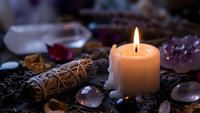 Burning candle surrounded by healing crystals, sage bundle and gemstones creating mystical spiritual atmosphere for meditation and energy cleansing rituals.