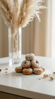 Freshly baked donuts dusted with powdered sugar arranged on white marble surface with dried pampas grass in glass vase creating warm minimalist kitchen scene.