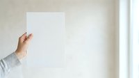 Close-up of a hand holding a blank white card or paper against a neutral wall background, suitable for mockups, presentations, and advertising campaigns.