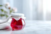 Homemade red berry jam in glass jar with fabric cover on marble table, bright kitchen setting with soft natural light.