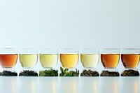 Different types of tea leaves and brewed tea in glass cups arranged in row showing variety of colors from light yellow to amber, with dry leaves displayed below.