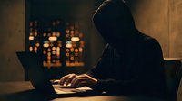 Anonymous hooded figure typing on laptop computer in dark room with city lights background for cybersecurity and hacking concepts.