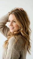 Young woman with long wavy blonde hair and fair complexion wearing a beige knit sweater, posing against white background with hand in hair, smiling at camera for portrait photography.