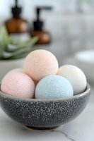 Pastel bath bombs in textured gray ceramic bowl on marble surface, with amber bottles and aloe plant in soft focus background. Spa and wellness composition.