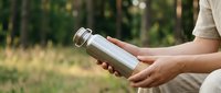 Woman holding stainless steel thermos bottle outdoors in forest setting for hiking camping and eco-friendly hydration lifestyle.