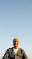 Senior man with gray hair and dark skin tone wearing beige cardigan over white shirt, photographed against clear blue sky during golden hour for portrait, lifestyle, and aging themes.