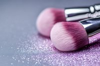 Pink makeup brushes with scattered glitter on gray and purple background, soft focus close-up shot showing professional cosmetic tools texture and details.