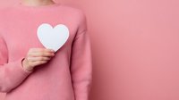 Person in pink sweater holding white heart shape against matching pink background for Valentine's Day, love concept, or romantic greeting card design.