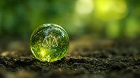 Glass sphere with scales of justice symbol resting on soil against blurred green forest background, representing environmental law and nature conservation.