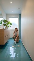 Woman in casual attire mopping teal floor in modern hallway with wooden vanity, potted plant, and natural light from window for cleaning and home maintenance concepts.