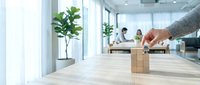 Cropped hand stacking wooden blocks in modern office with colleagues working at desk and green plant in background, representing teamwork, strategy and business growth.