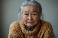 Portrait of senior Asian woman with gentle smile wearing cozy knit sweater against blurred background, conveying wisdom and contentment in natural lighting.