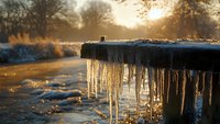 Glowing winter sunrise illuminates frozen icicles hanging from wooden fence, creating magical morning atmosphere with golden light and frost covered landscape.