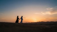 Two women in traditional long dresses hold hands while walking across desert sand during golden hour sunset, silhouetted against warm orange and pink sky with mountains in distance.