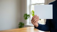 Person holding blank white card or paper in office environment with natural light and green plants in background for mockup or advertising purposes.