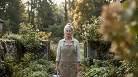 Mature Caucasian woman in linen apron standing in lush garden, holding watering can, surrounded by green plants and stone wall.