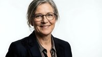Mature woman with gray hair and eyeglasses wearing black blazer and dark shirt, smiling at camera against white background for corporate portraits.