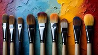 Professional makeup brushes arranged in row against colorful textured background showing blue, yellow and red paint strokes. Artistic cosmetic tools composition.