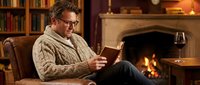 Middle-aged Caucasian man in cozy sweater reading book by fireplace in home library with wine glass, relaxing evening atmosphere for lifestyle content.