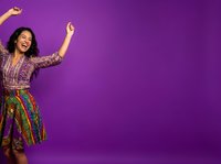 Young woman with long dark hair and South Asian ethnicity wearing colorful patterned shirt and skirt, dancing with arms raised against vibrant purple background. Suitable for lifestyle, celebration, and happiness concepts.