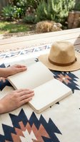 Person reading blank book outdoors on patterned rug with straw hat, hands turning pages in sunny garden setting for relaxation and leisure activities.