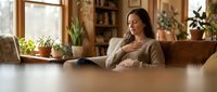 Pregnant woman relaxing on sofa at home reading book in cozy living room with plants and natural light streaming through windows.