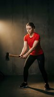Athletic young woman in red shirt swinging axe in dramatic gym lighting for strength training workout and functional fitness exercise.