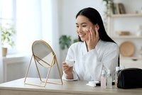 Happy Asian woman applying skincare cream while looking in mirror at home, morning beauty routine with cosmetic products on wooden table.