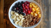 Fresh fruit and granola bowl with banana slices, blackberries, raspberries, mango chunks, almonds and crunchy granola on rustic wooden background.