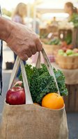 Close-up of mature male hand holding canvas tote bag filled with fresh produce including kale, red apple, and orange at farmers market or outdoor grocery shopping location.