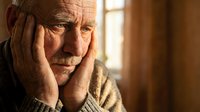Senior Caucasian man with hands on face showing stress, depression or headache in warm indoor lighting. Mental health and elderly care concept.