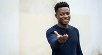 Young African American man in navy sweater making welcoming gesture with outstretched hand, smiling warmly against neutral background for business content.