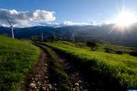 Wind turbines on green mountain ridge with dramatic sunburst through clouds, dirt hiking trail leads through spring meadow landscape at sunset. Renewable energy concept.