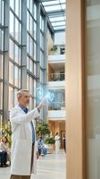 Senior Caucasian male doctor in white coat interacting with holographic medical interface in modern hospital atrium with glass architecture and natural light.