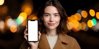 Young Caucasian woman with brown hair showing blank smartphone screen against blurred city lights at night, wearing beige coat.