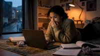 Young mixed-race woman lying on floor, anxiously studying on laptop at night, surrounded by books and crumpled papers in cozy room.