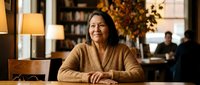 Mature Asian woman smiling warmly while sitting at wooden table in cozy cafe or restaurant with autumn decor and soft lighting for lifestyle content.