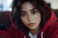 Young Asian woman in red hoodie looking directly at camera with intense gaze, natural makeup, and tousled dark hair against blurred background.