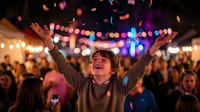 Young woman celebrating with raised arms at outdoor festival with colorful lights and confetti falling around crowd of people enjoying live music event.